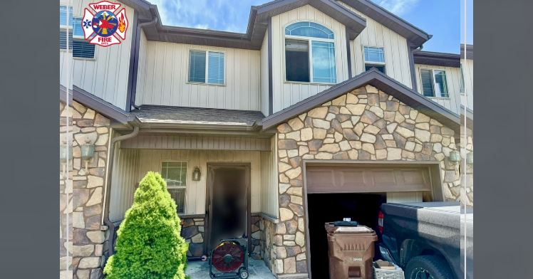 Two story suburban home with stone accents damaged front entry open garage a fan and trash cans in the driveway Weber Fire emblem in the top left corner