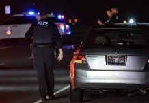Nighttime police stop: rear of a silver Honda with flashing blue and red lights in the background and officers nearby on a dark road.