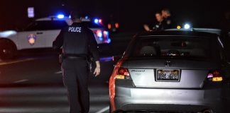 Nighttime police stop: rear of a silver Honda with flashing blue and red lights in the background and officers nearby on a dark road.