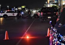 Nighttime city street with police cars, flashing lights, and orange traffic cones at a roadblock. Officers stand near the vehicles amid scattered streetlights and storefronts in the background.