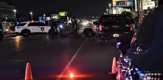 Nighttime city street with police cars, flashing lights, and orange traffic cones at a roadblock. Officers stand near the vehicles amid scattered streetlights and storefronts in the background.