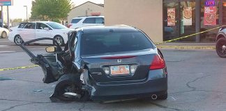 Blue Honda Accord with extensive driver-side damage after a crash; driver's door open and debris on pavement with caution tape in background of a storefront lot.