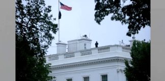 White House rooftop with an American flag flying, a person standing near the railing, framed by trees against a pale sky.