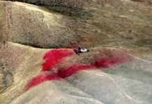 Rugged hillside with a small vehicle on a dusty slope and a large red stain on the ground ahead.] ,