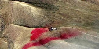 Rugged hillside with a small vehicle on a dusty slope and a large red stain on the ground ahead.] ,