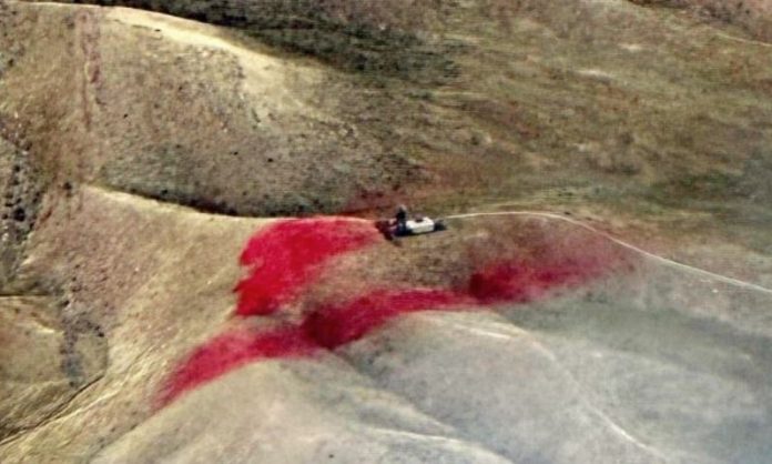Rugged hillside with a small vehicle on a dusty slope and a large red stain on the ground ahead.] ,