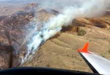 In-flight view of a wings level aircraft with orange wingtip over dry hills, smoke rising from a hillside fire below.