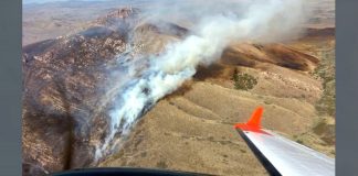 In-flight view of a wings level aircraft with orange wingtip over dry hills, smoke rising from a hillside fire below.