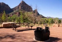 Picnic area in a desert park featuring a round charcoal grill, a wooden picnic table, and sparse trees with rugged mountains in the background under a clear blue sky