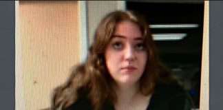 Young woman with long brown hair wearing a black top, looking slightly to the left, indoors in a dim background.
