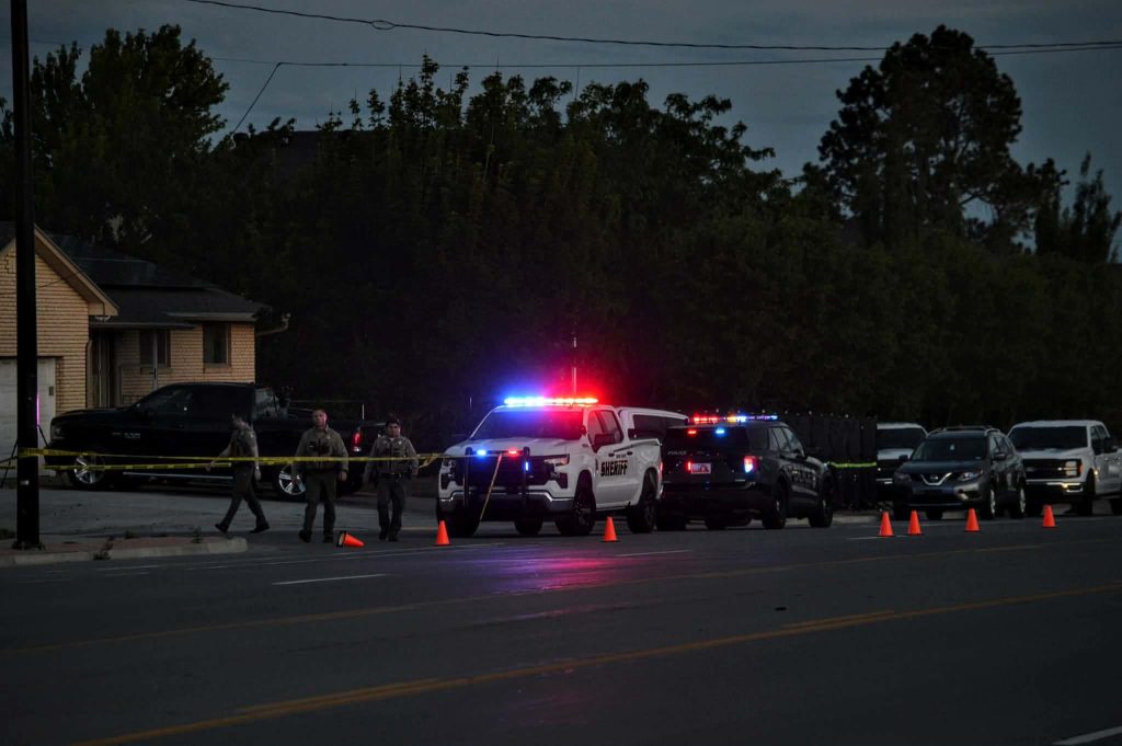Nighttime street scene with police and sheriff vehicles, flashing red and blue lights, and yellow caution tape cordoning off the area outside a residential building.