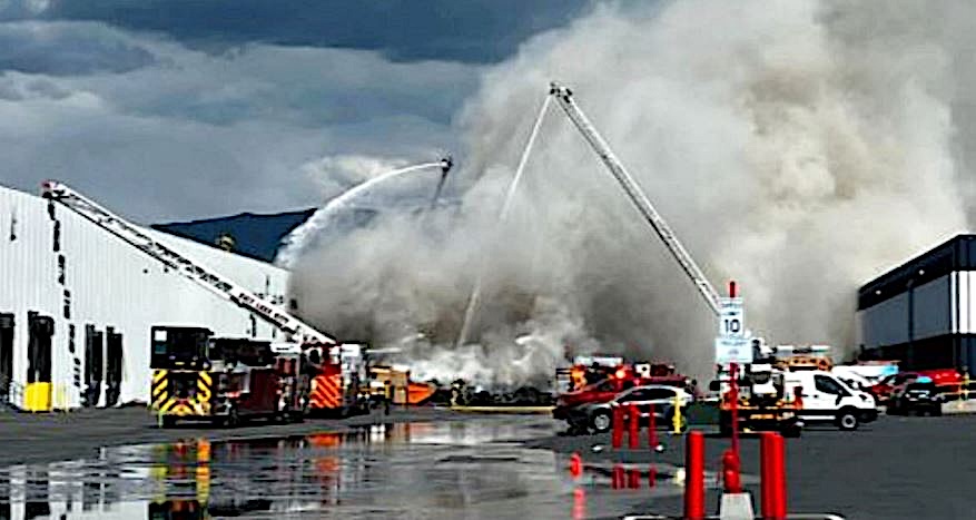 Firefighters battle a large warehouse fire with extended ladders spraying water amid thick smoke.