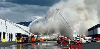 Firefighters battle a large blaze at an industrial warehouse with water streams and thick smoke rising above the building.