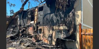 Severely fire-damaged house with blackened walls, charred debris scattered in the front yard, and a wooden fence to the right.