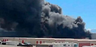 Large dark smoke plume rising from a warehouse district, with trucks and storage buildings in the foreground.