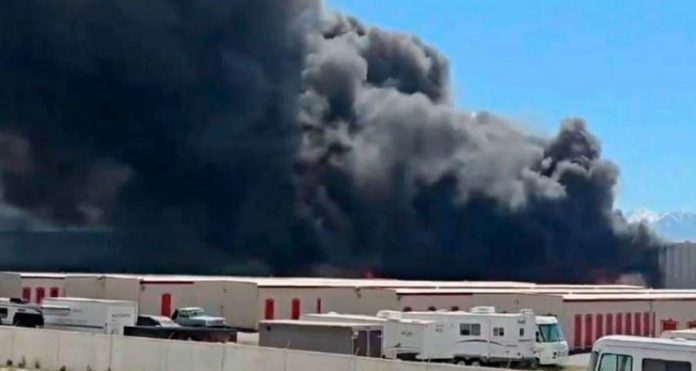 Large dark smoke plume rising from a warehouse district, with trucks and storage buildings in the foreground.