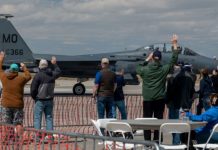 Military fighter jet on a runway with spectators behind a safety barrier waving at the plane.