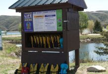 Life jacket loaner station by a lake, with numerous yellow life jackets on racks under a wooden shelter.