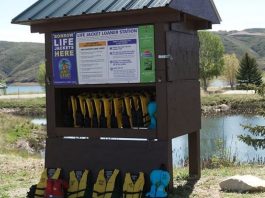 Life jacket loaner station by a lake, with numerous yellow life jackets on racks under a wooden shelter.