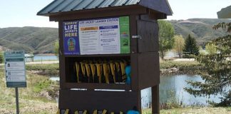 Life jacket loaner station by a lake, with numerous yellow life jackets on racks under a wooden shelter.