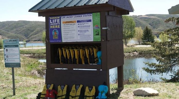 Life jacket loaner station by a lake, with numerous yellow life jackets on racks under a wooden shelter.