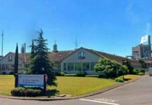 Single-story medical facility with a sloped roof, a well-kept lawn, and trees, flagpoles with flags, and a driveway; a sign is visible on the left under a clear blue sky.