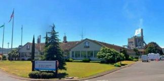 Single-story medical facility with a sloped roof, a well-kept lawn, and trees, flagpoles with flags, and a driveway; a sign is visible on the left under a clear blue sky.