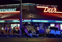 Overturned white car on the sidewalk in front of a neon-lit diner at night, with a crowd watching the crash scene.