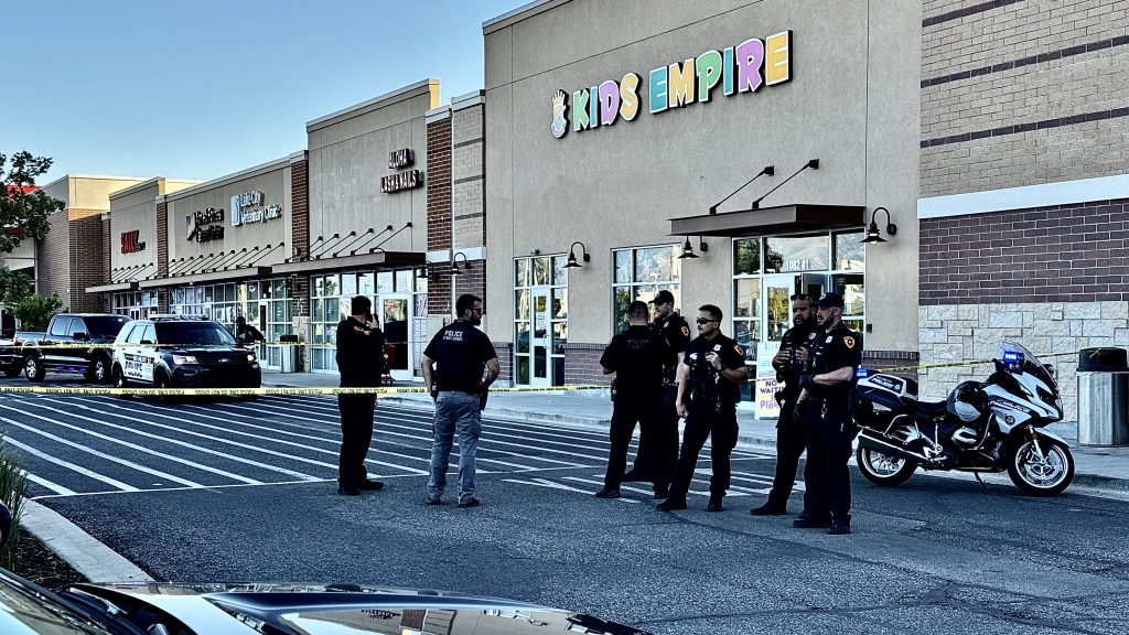 Salt Lake City shooting | Gephardt Daily Group of police officers standing behind yellow caution tape outside a strip mall with storefronts including a colorful'KIDS EMPIRE' sign; police cars and a motorcycle are parked nearby.