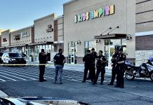 Group of police officers standing behind yellow caution tape outside a strip mall with storefronts including a colorful 'KIDS EMPIRE' sign; police cars and a motorcycle are parked nearby.