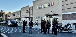 Group of police officers standing behind yellow caution tape outside a strip mall with storefronts including a colorful 'KIDS EMPIRE' sign; police cars and a motorcycle are parked nearby.
