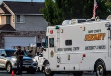 Davis County Sheriff mobile command center emergency vehicle parked on a suburban street, a deputy speaks on a radio beside a silver SUV.