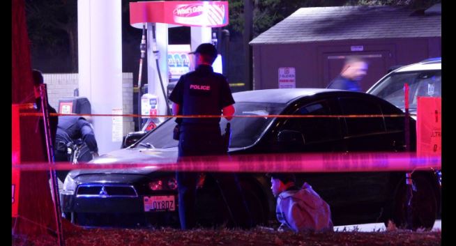 Police officer at a gas station crime scene near a dark sedan, pink tape in the foreground, with a person sitting on the ground beside the car (evening scene).