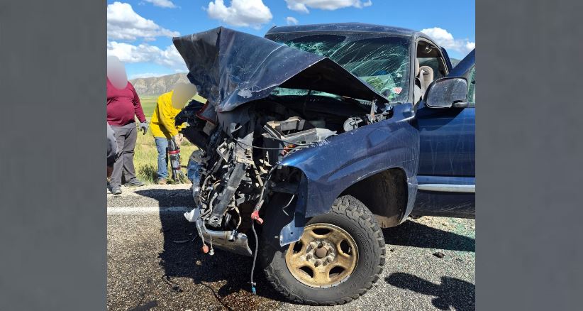 Blue pickup truck with severe front end damage after a collision on a sunny road bystanders nearby