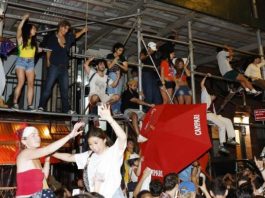 Crowd dancing on metal scaffolding above a street party at night, with a red umbrella and people cheering.