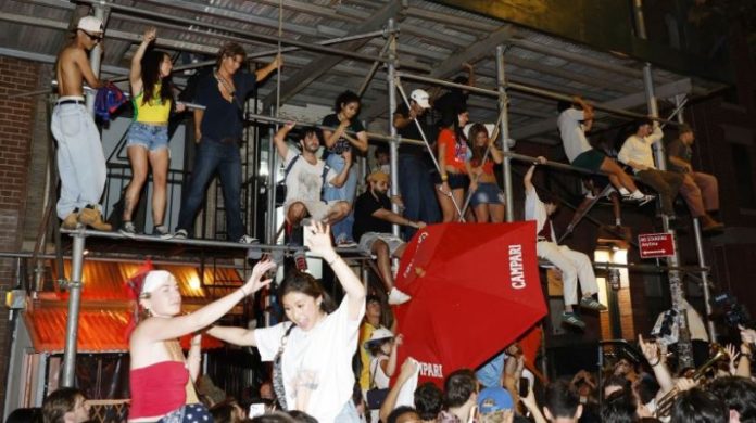 Crowd dancing on metal scaffolding above a street party at night, with a red umbrella and people cheering.