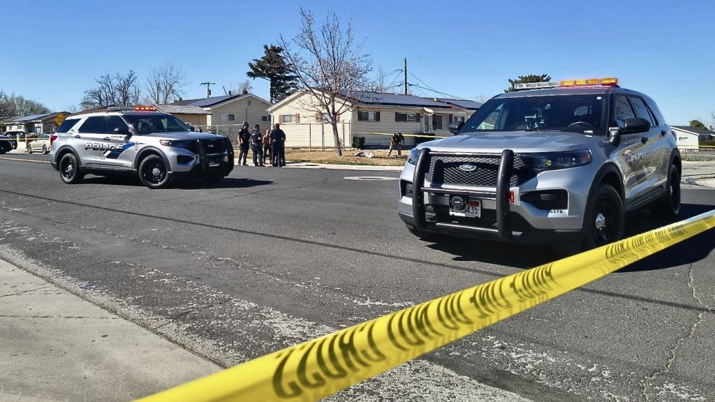 Police SUVs with flashing lights on a residential street behind yellow caution tape; officers gather near a house under a clear blue sky.