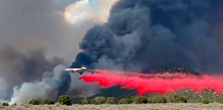 Plane drops red fire retardant over a brush fire, with dark smoke plumes rising in the background.