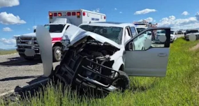 White pickup with severe front-end damage sits off the road in tall grass; driver door open and an ambulance behind.