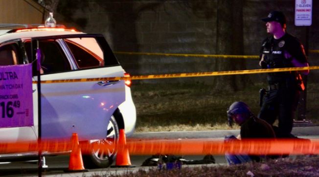Police scene at night: a white SUV with open door, orange traffic cones, yellow tape, and an officer guarding a kneeling person near the ground.