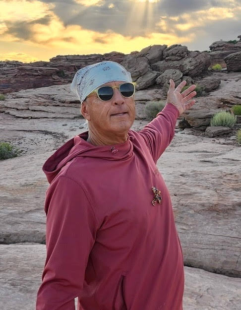 25-andy-lewis-danny-kregle | Gephardt Daily Man in a maroon hoodie and light bandana, wearing sunglasses, stands on rocky terrain with arms outstretched at sunset.