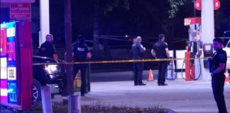 Police officers stand behind yellow caution tape at a dimly lit gas station scene, documenting evidence at night.