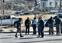 A group of people standing behind yellow caution tape in a parking lot, some wearing jackets, with cars and suburban houses in the background.