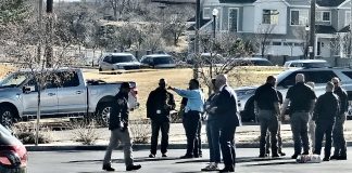 A group of people standing behind yellow caution tape in a parking lot, some wearing jackets, with cars and suburban houses in the background.