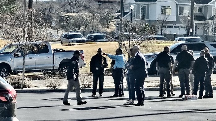 A group of people standing behind yellow caution tape in a parking lot, some wearing jackets, with cars and suburban houses in the background.