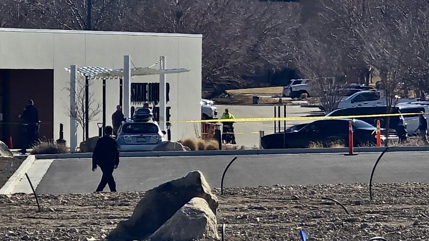 Law enforcement at an outdoor scene outside a white building, with yellow caution tape and several vehicles nearby.