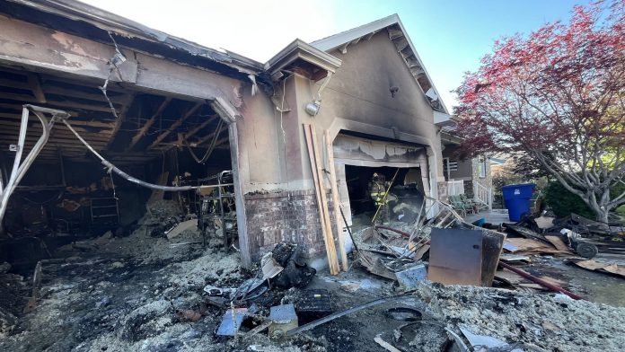 Charred, partially collapsed house with debris in the yard and a firefighter near the garage.