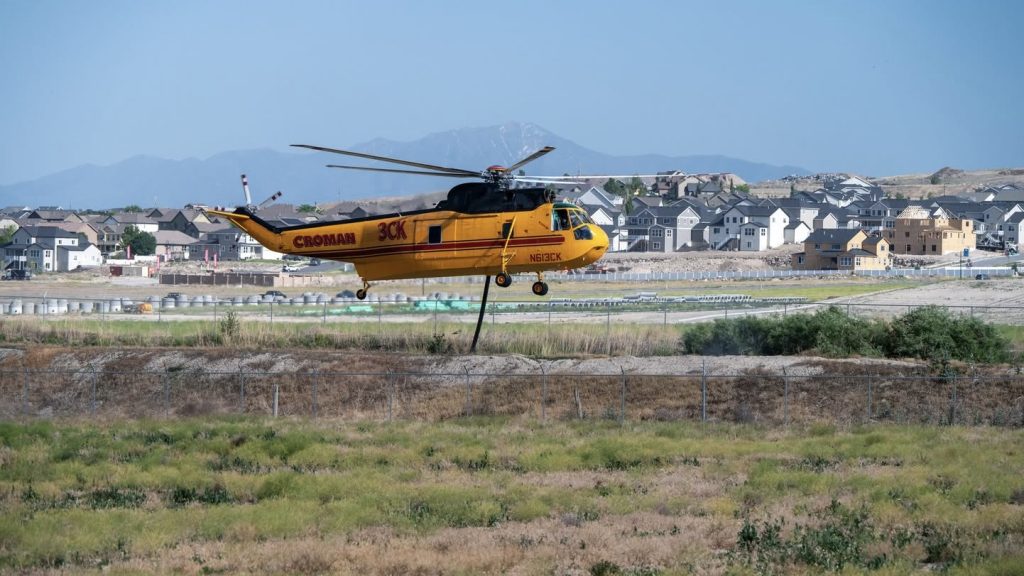 Yellow helicopter labeled 'CROMAN 3CK' lifting off over a grassy field with a fence and a suburban neighborhood beyond.