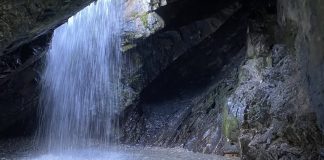 Waterfall spilling from a rocky cave ceiling into a small pool among dark jagged walls.