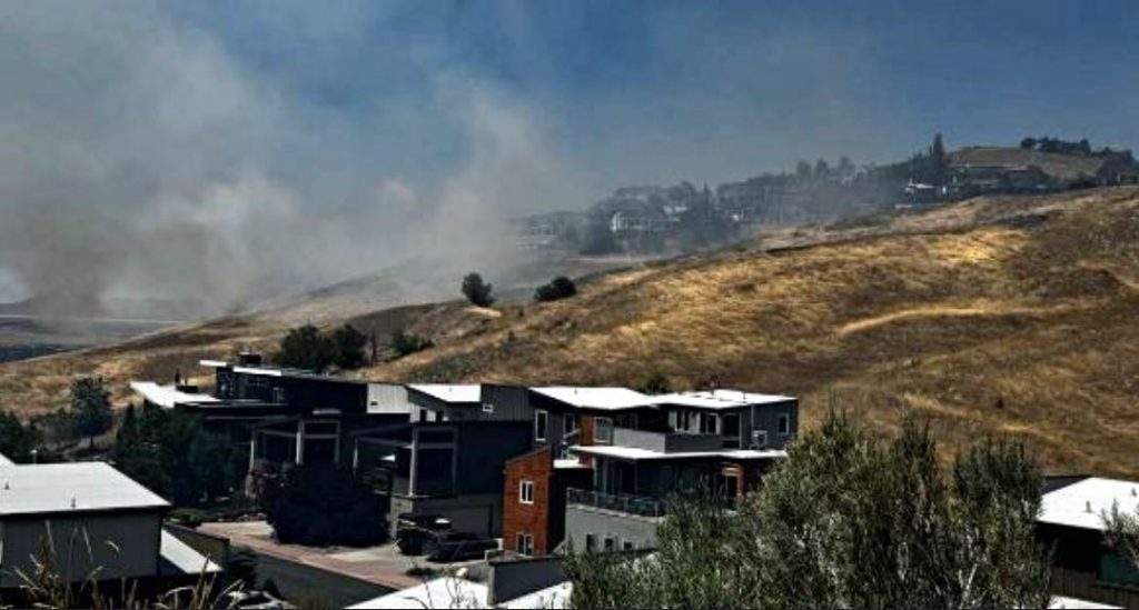 Hillside neighborhood with modern houses and smoke rising in the distance, indicating a wildfire.
